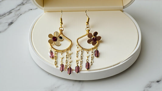 Gold earrings with floral designs and red beads on a white jewelry box against a marble background