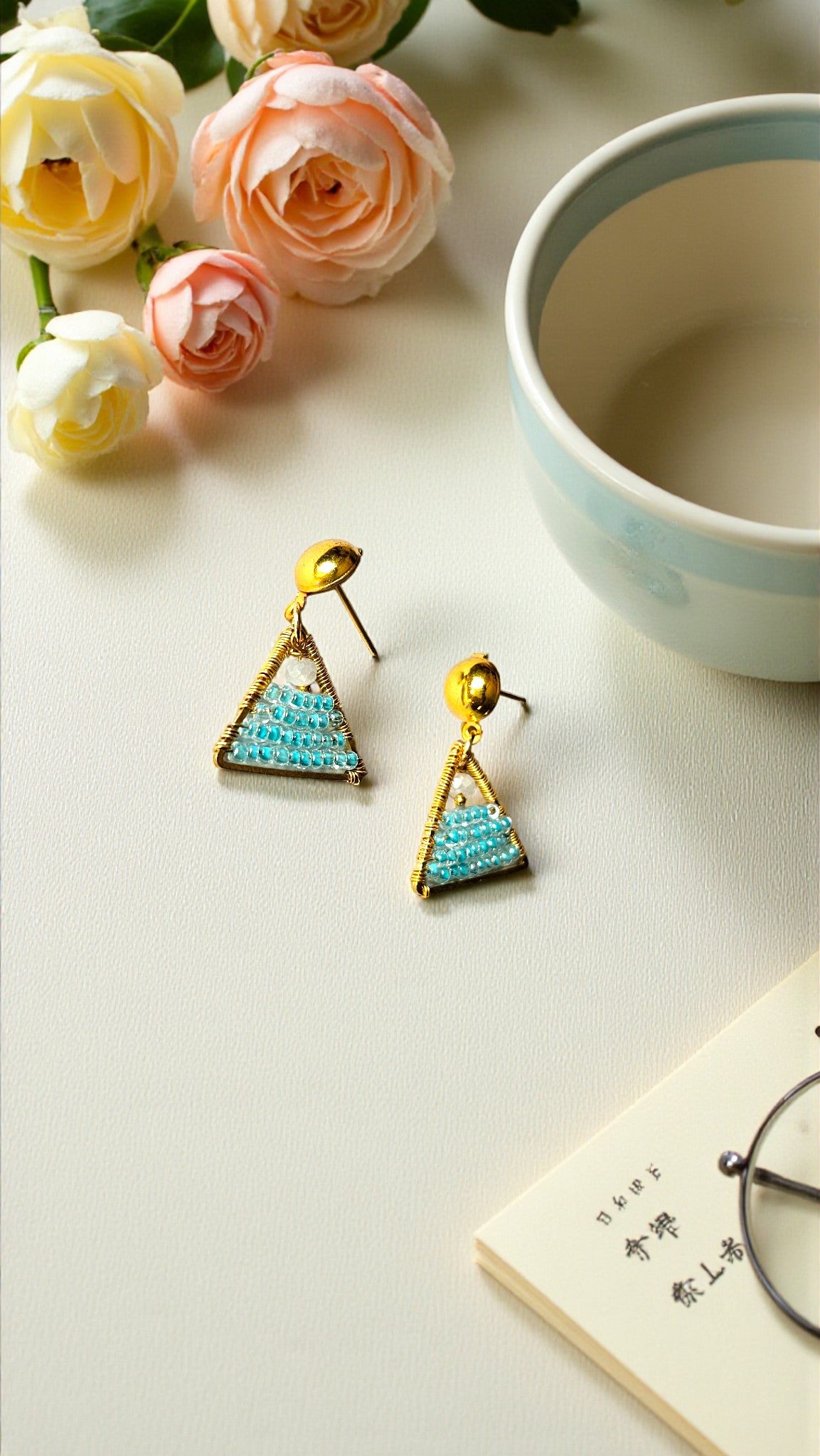 Triangle-shaped earrings with turquoise stones on a light surface with flowers and a cup.