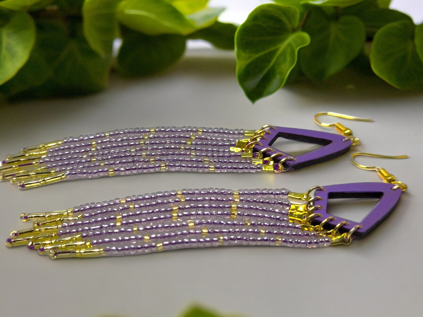 Purple and gold beaded earrings on a light surface with green leaves in the background