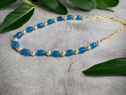 Affordable beaded necklace with blue and pearl beads and gold chain on a stone surface with green leaves