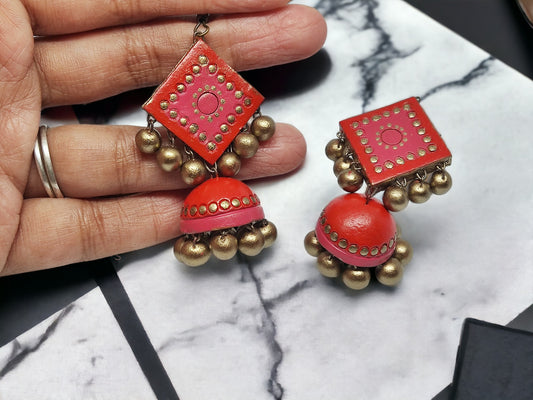 Hand holding handmade Bhairavi Terracotta Jhumka earrings in red with gold bead accents from Anusangi Collections