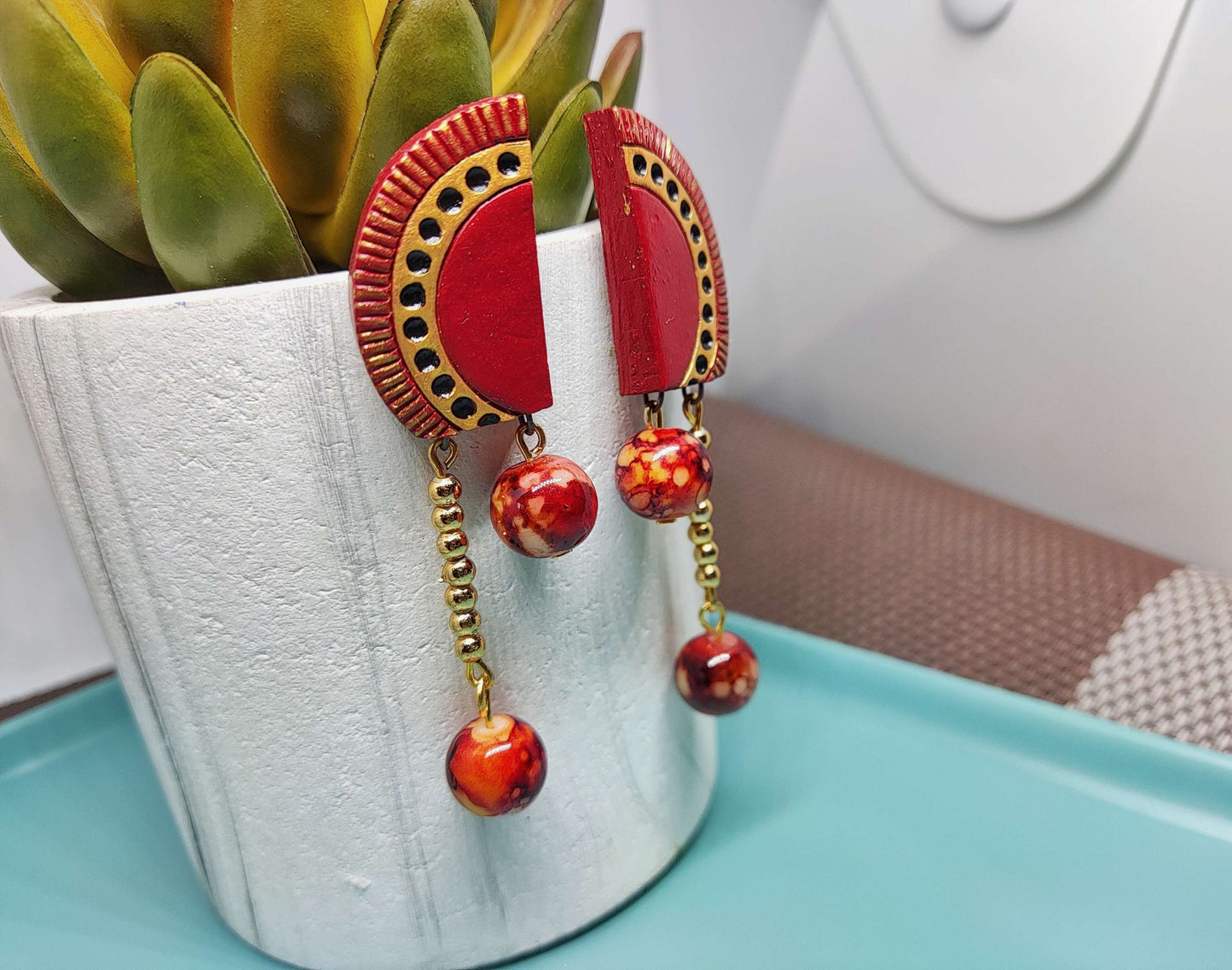 Anusangi Collections red and gold statement terracotta earrings with dangling beads displayed on a white pot