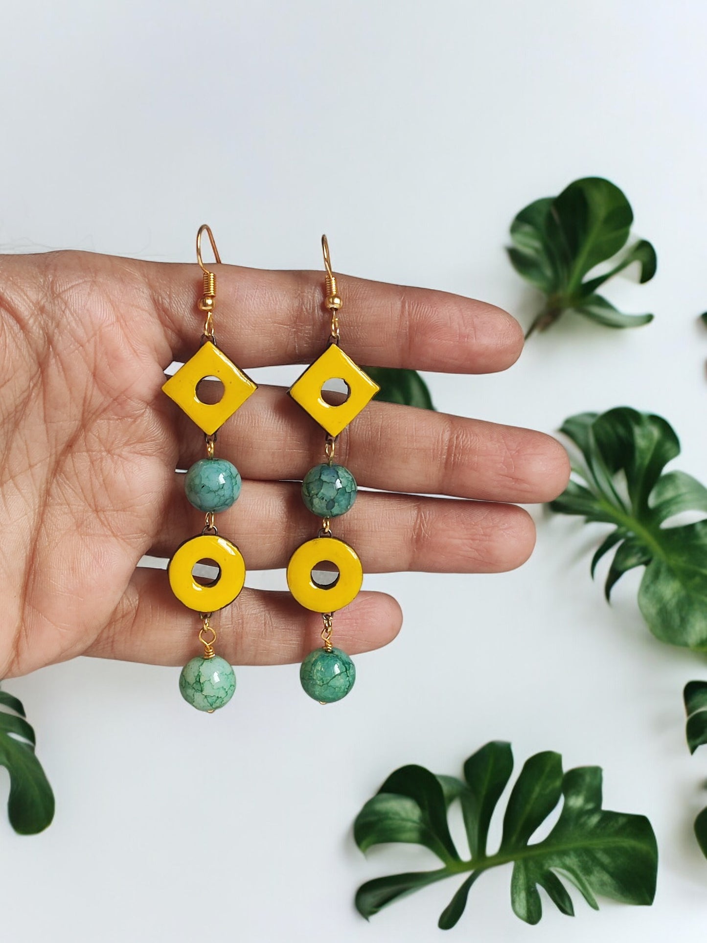 Yellow and green dangle statement terracotta earrings held in hand with leafy background