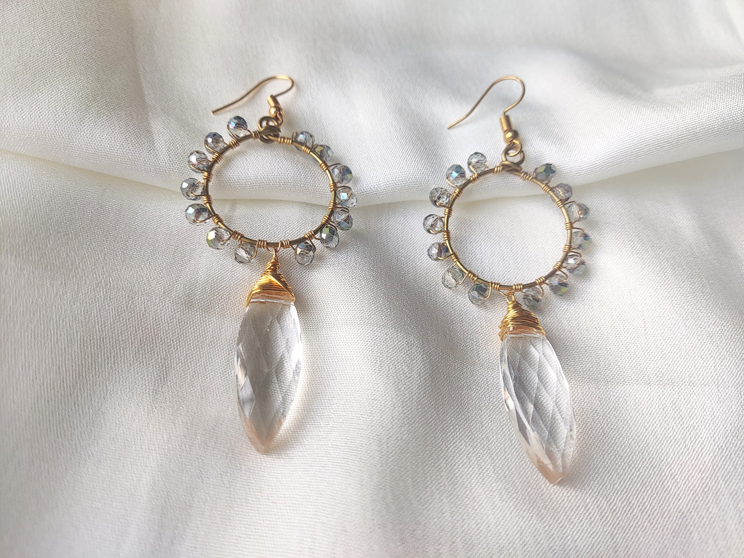 Elegant beaded earrings Collections featuring gold hoops with clear crystal beads and teardrop pendants on white fabric