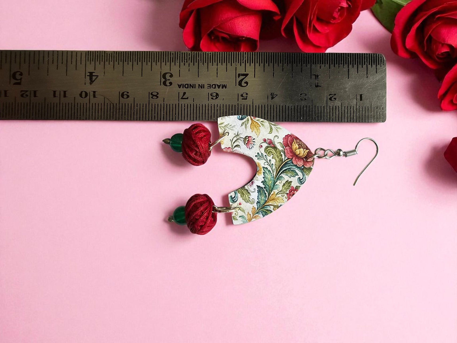 Floral patterned earrings with red floral charms on a pink background with a ruler for scale.