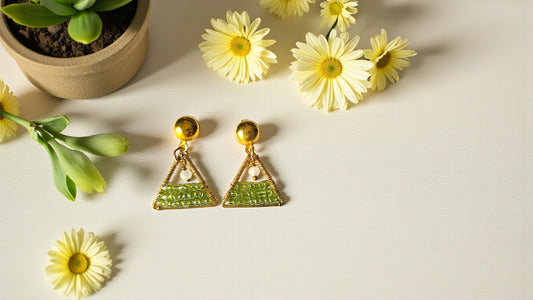 Triangular earrings with green gemstones on a light surface with flowers and a plant.