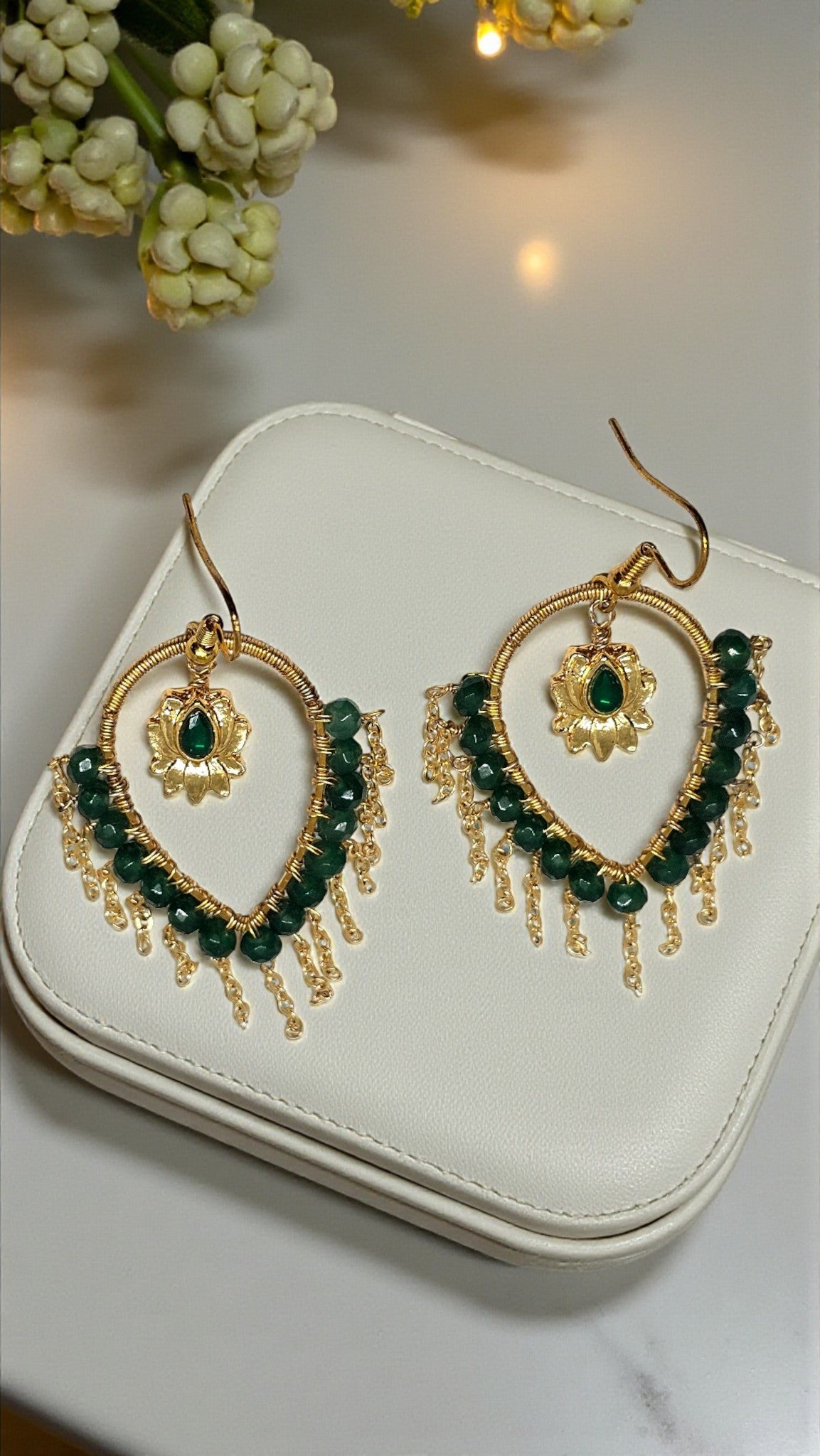 Gold and green beaded earrings on a white jewelry case with flowers in the background