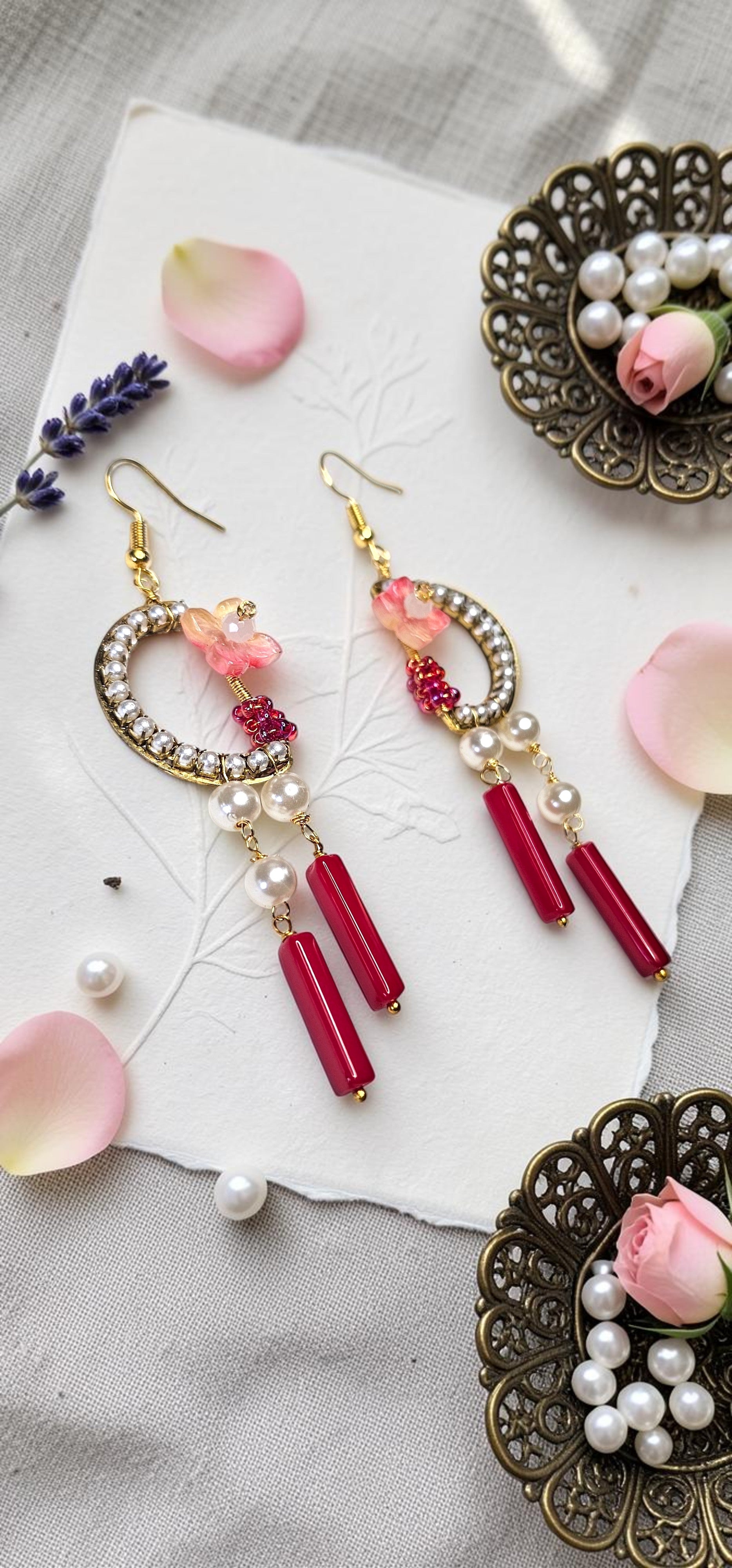 A pair of gold-tone dangle earrings with rhinestone-embellished crescent hoops, pink floral charms, white faux pearls, and dangling red cylindrical beads, styled on a white textured card with rose petals and decorative metal dishes.