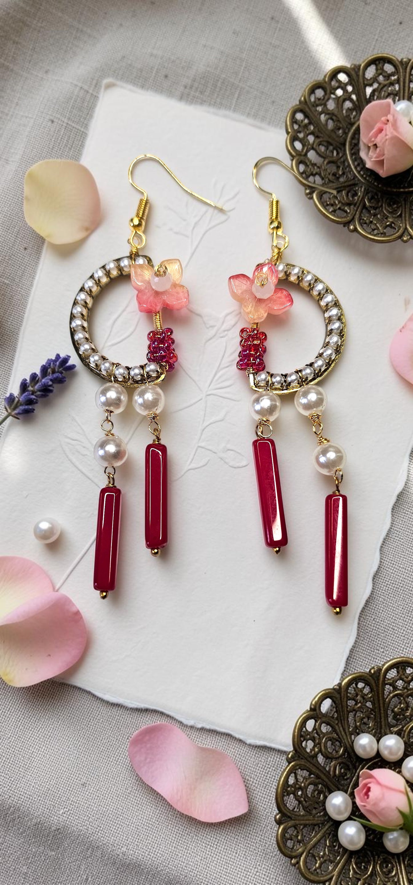 A pair of gold-tone hoop earrings with pink floral charms, white pearl-like beads, and red cylindrical drops, arranged on a white paper sheet with pink petals and decorative metal pieces around them.