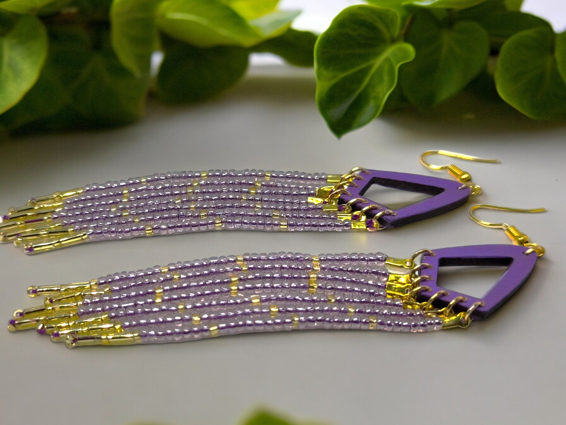 Purple and gold beaded earrings on a light surface with green leaves in the background