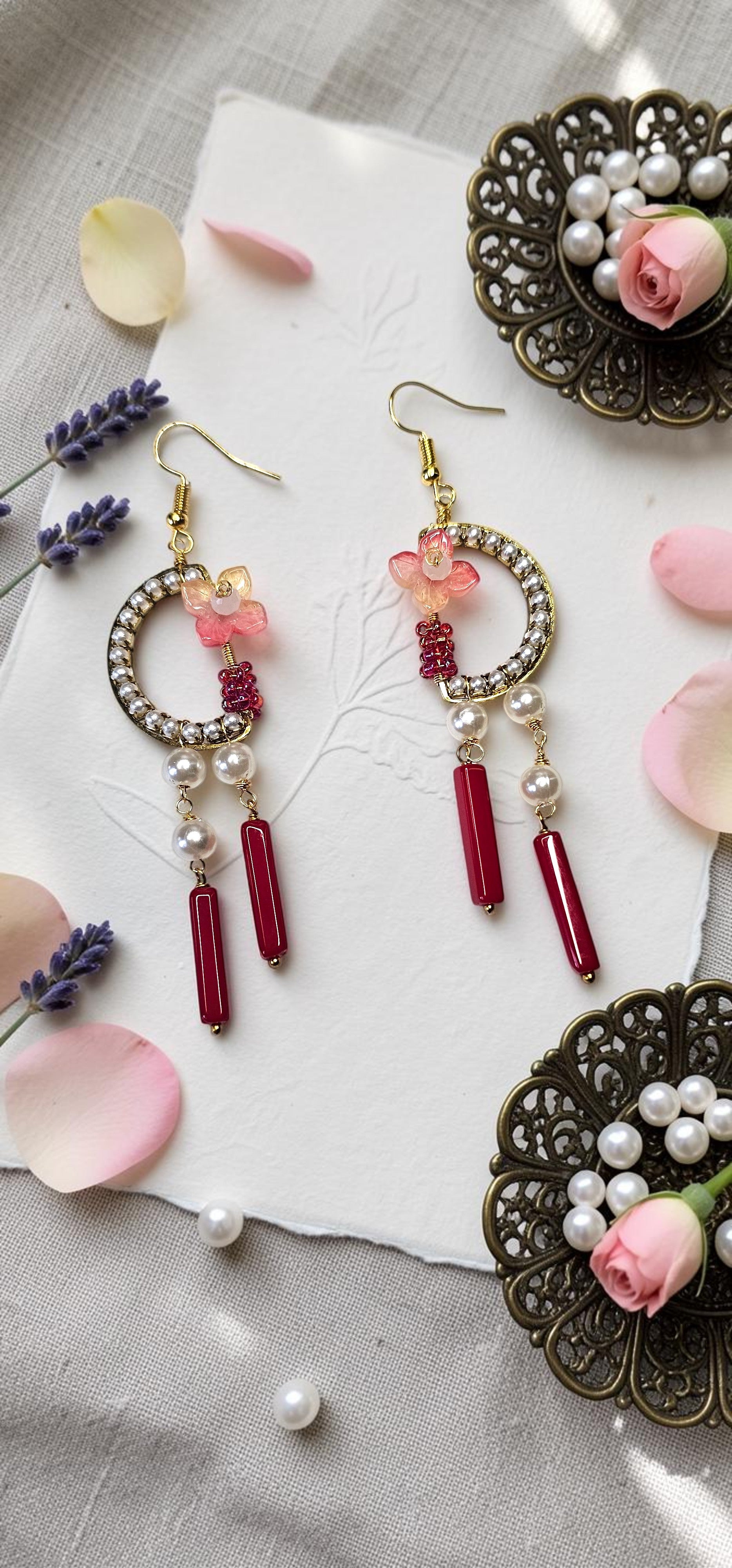 A pair of gold-toned dangle earrings with circular crystal-rimmed hoops, pink floral charms, white pearl drops, and dark red cylindrical beads, styled on a white paper sheet with pink petals and decorative metal rose holders.
