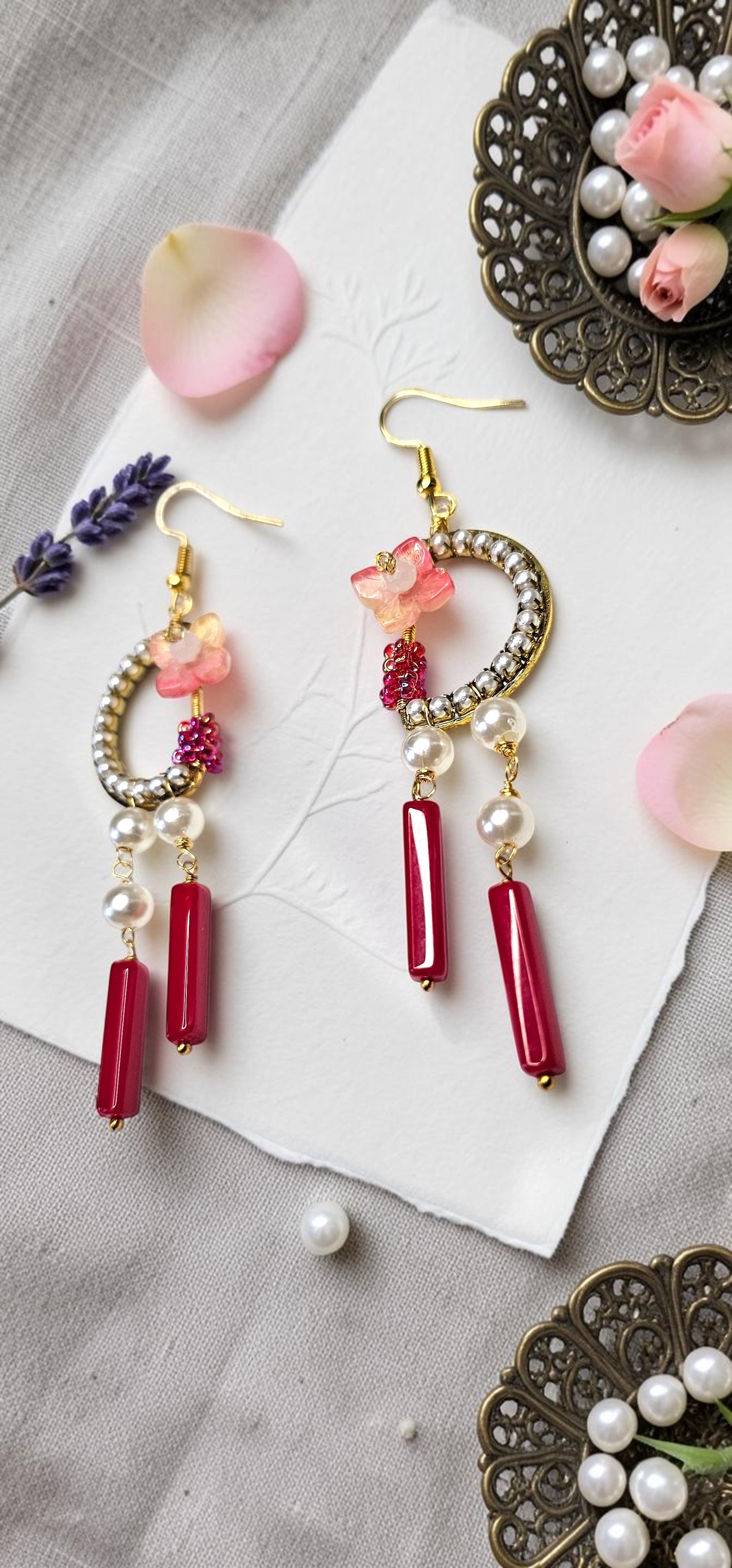 A pair of gold-tone dangle earrings with pink floral charms, white pearl beads, and red cylindrical drops, styled on a textured gray surface with decorative props.