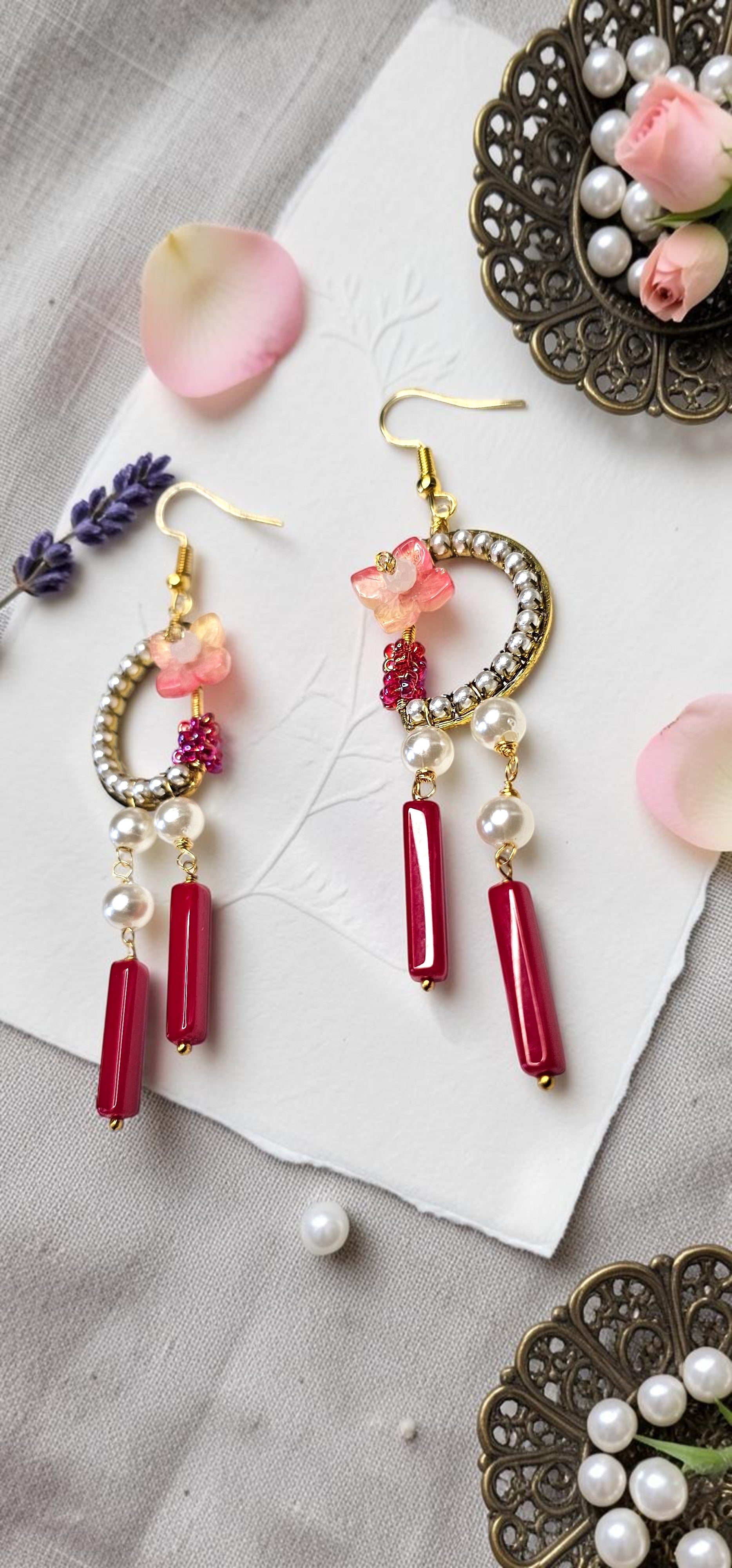 A pair of gold-tone dangle earrings with pink floral charms, white pearl beads, and red cylindrical drops, styled on a textured gray surface with decorative props.