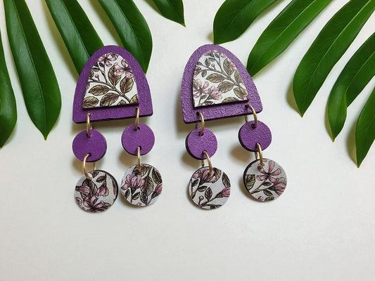 Pair of purple floral earrings on a white background with green leaves.