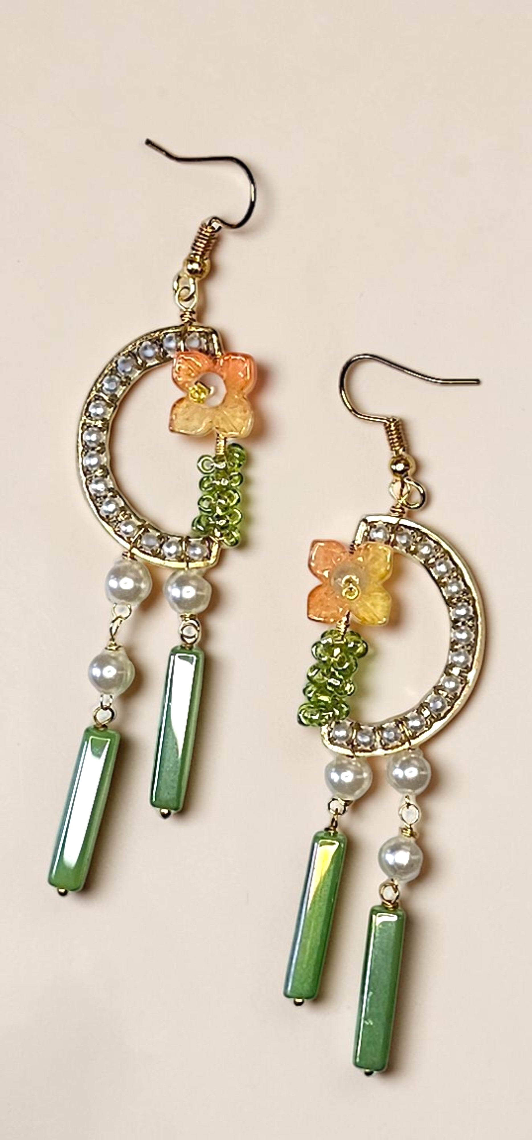A pair of gold-tone dangle earrings featuring crescent-shaped hoops set with clear rhinestones, small orange flower charms, green beaded details, white pearl beads, and elongated green baroque beads, displayed on a light beige background.